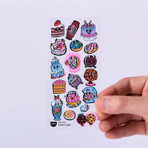 A hand holds a sheet of Criossant - Fabulous Stickers (Copy) featuring colourful cartoon desserts like cake slices, doughnuts, milkshakes, biscuits, and fun phrases such as "Born to Be Sweet," set against a light purple background. Gate21.ee