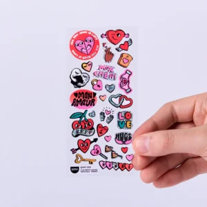 A hand holds the Letter - Fabulous Stickers, featuring hearts, cherries, shoes, and playful text like Mon Amour, LOVE, HUGS, and AMOR. These stickers add extra charm to the white background. Gate21.ee