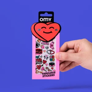 A hand holds a pink pack of "Letter - Fabulous Stickers (Copy)" featuring colourful heart-themed stickers and playful earring stickers by OMY, with a smiling red heart on the packaging top, against a solid blue background. Gate21.ee