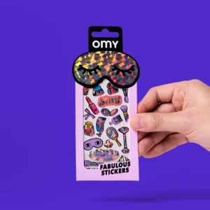 A hand holds a pack of Heart - Fabulous Stickers (Copy), featuring colourful makeup items, accessories, sticker earrings, and “Slay” on the front. The packaging has a shiny black sleep mask at the top against a purple background. Gate21.ee