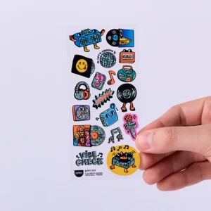 A hand holds a sheet of Lashes - Fabulous Stickers, showcasing colourful cartoon music-themed objects like cassette tapes, records, a boombox, speakers, and OMY sticker earrings with "Vibe Check" at the bottom. Gate21.ee