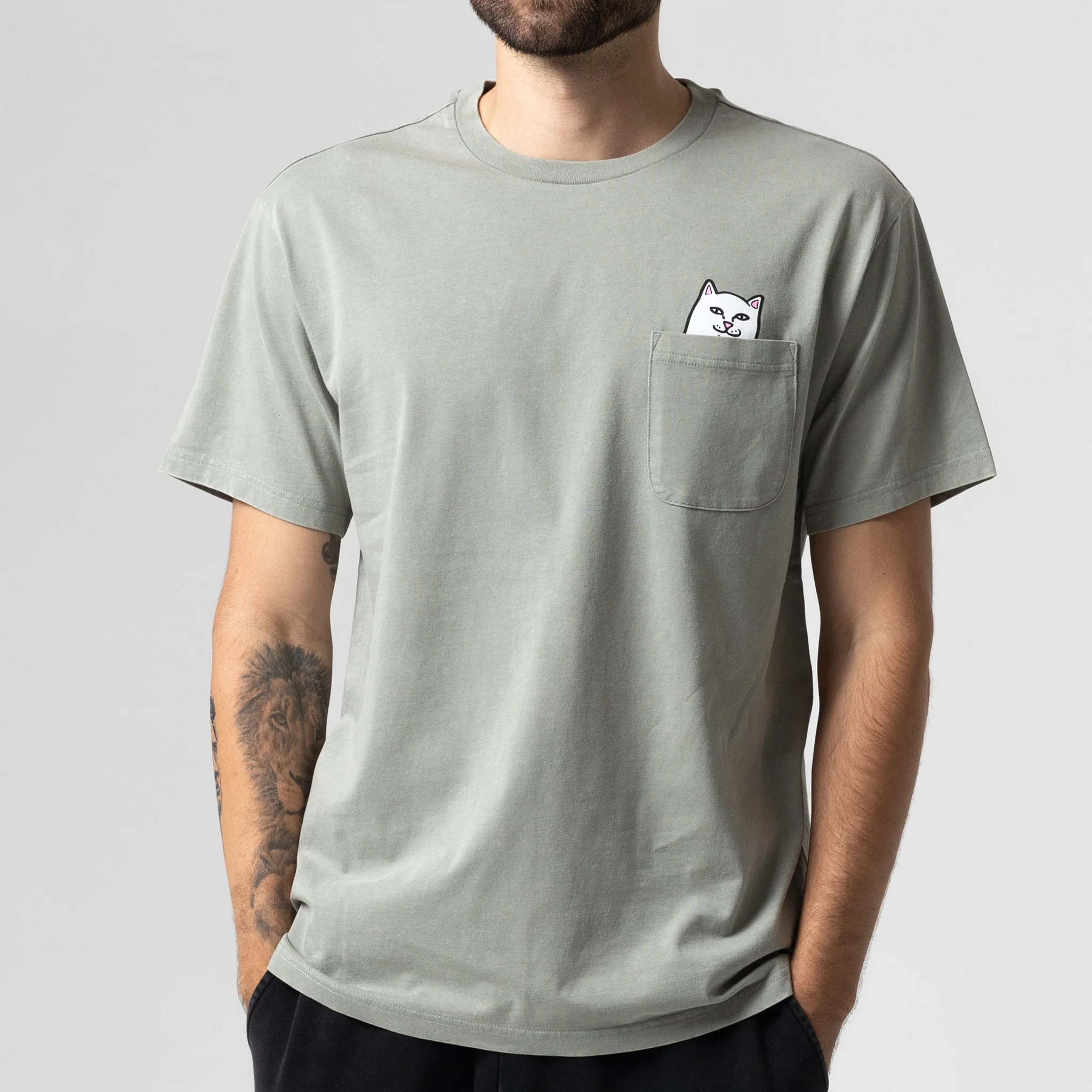 A person wears the Lord Nermal Pocket Tee in brown vintage wash—a T-shirt featuring an illustrated white cat peeking from the chest pocket. The person has a short beard and visible tattoos on one forearm. Gate21.ee
