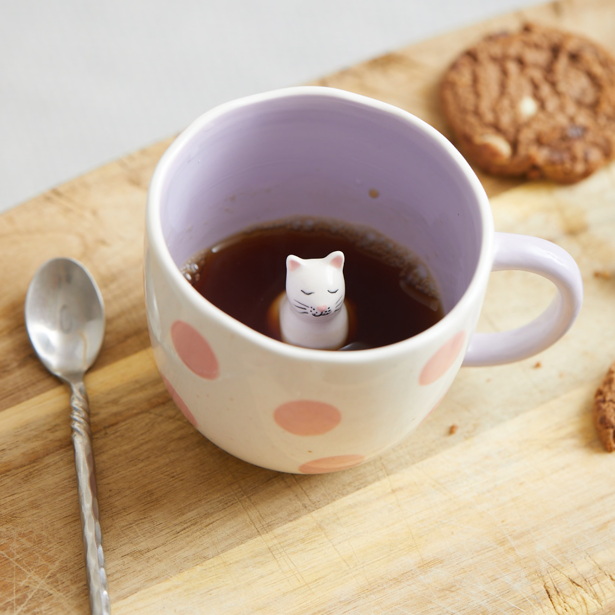 A Surprise Dog - Cup (Copy) with a light purple finish and pink polka dots sits on a wooden surface, filled with tea or coffee. Inside the cup is a ceramic dog figurine, with a metal spoon and two biscuits nearby. Gate21.ee
