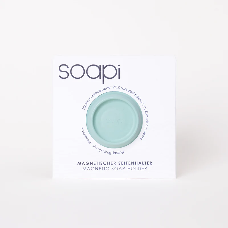 A round, yellow Soapi magnetic soap holder is shown in white packaging with the brand name and product details in English and German, highlighting that it’s made with about 90% recycled fishing nets. Gate21.ee