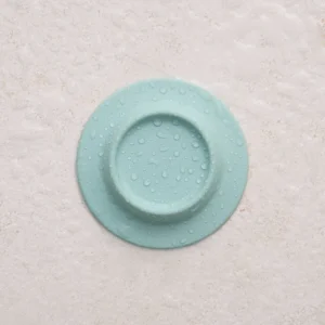 A round, yellow Soapi silicone drain cover is placed on a textured, off-white background. Gate21.ee