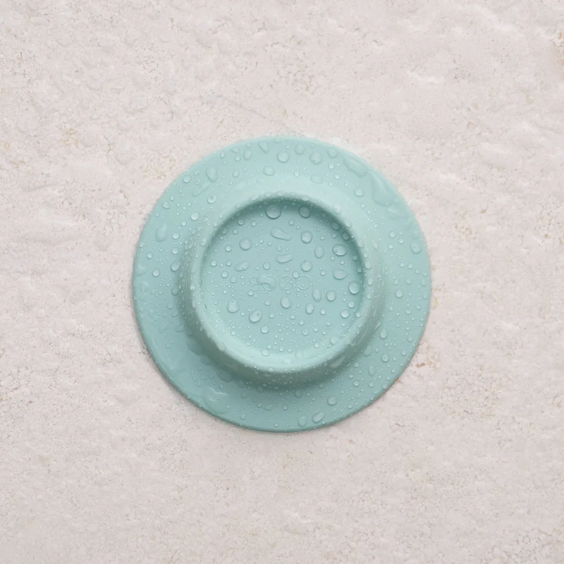 A round, yellow Soapi silicone drain cover is placed on a textured, off-white background. Gate21.ee