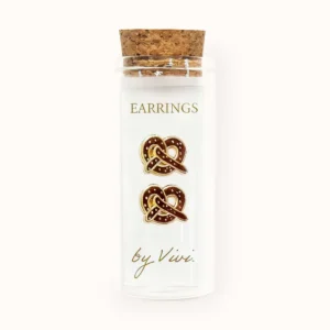 A clear glass phial with a cork lid holds a card labelled "Brezels by Vivi," displaying gold and brown pretzel-shaped earrings with white accents. Gate21.ee