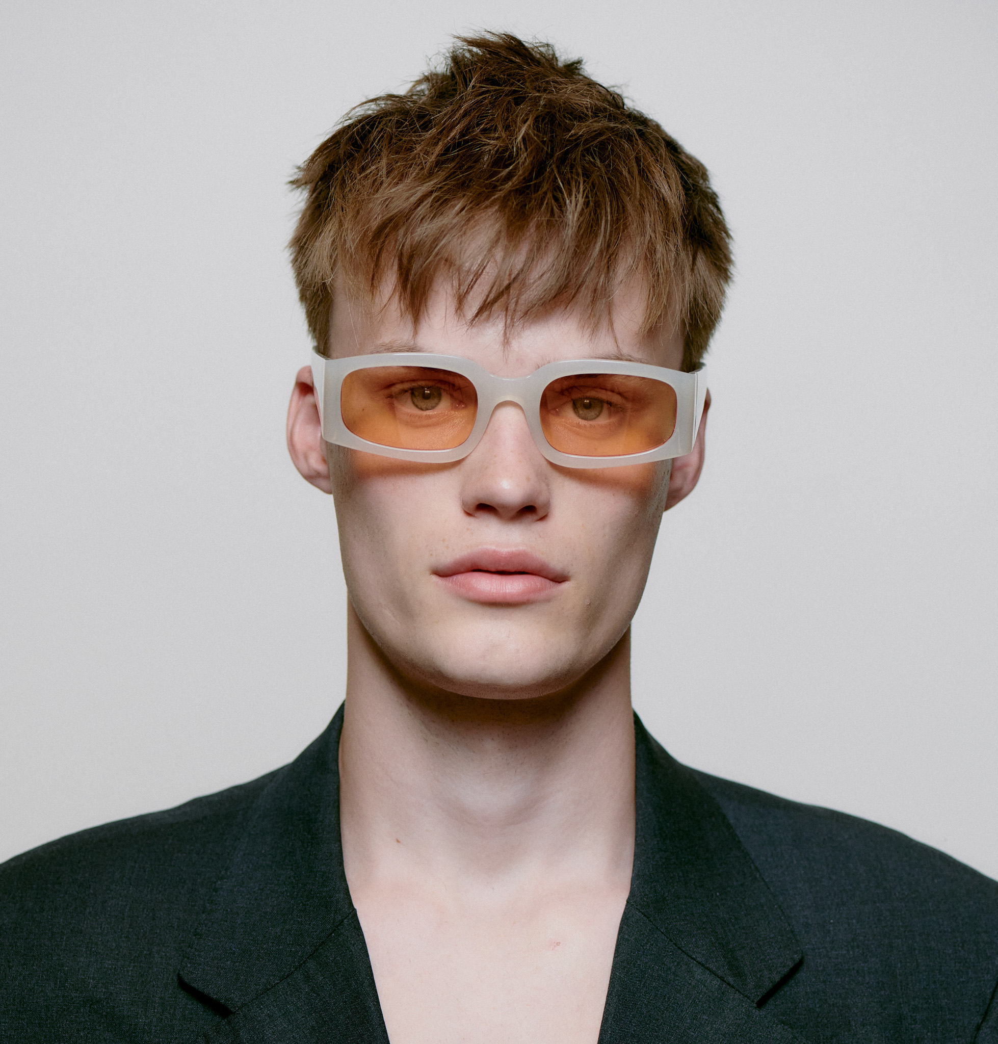 A person with short, tousled brown hair wears ALEX - Cream Bone white-framed, orange-tinted rectangular sunglasses and a dark blazer, looking directly at the camera against a plain light background. Gate21.ee