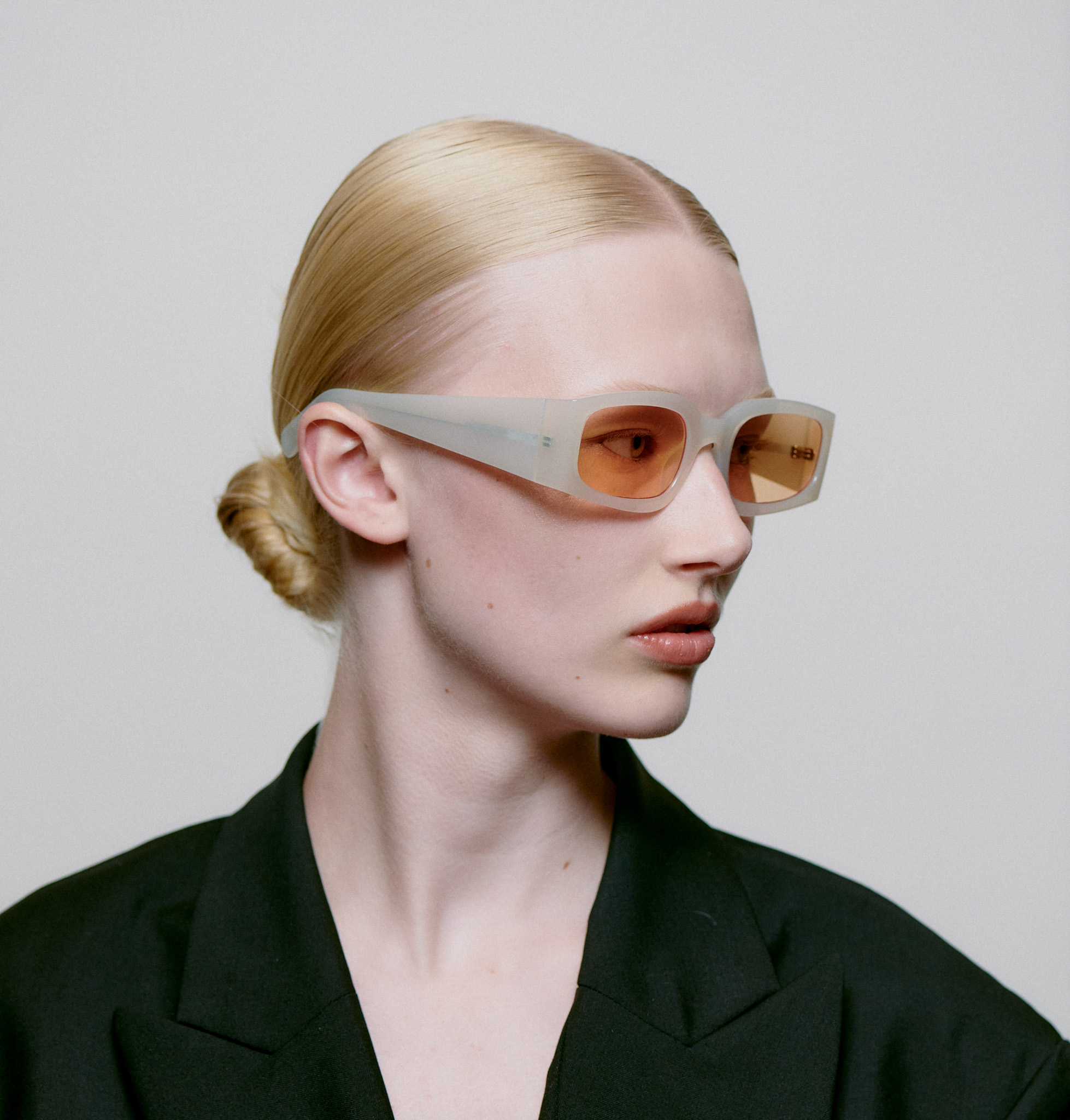 A person with blond hair in a low bun wears large, translucent cream-beige ALEX - Cream Bone sunglasses by A. Kjærbede with amber lenses and a black jacket, looking left against a plain light background. Gate21.ee