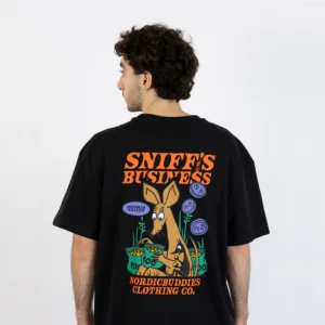 A person faces away, wearing the Moomin Heavy Ts - Sniff's Business featuring a colourful cartoon dog, plants, and the text "SNIFF'S BUSINESS" and "NORDICBUDDIES CLOTHING CO." printed on the back. Gate21.ee