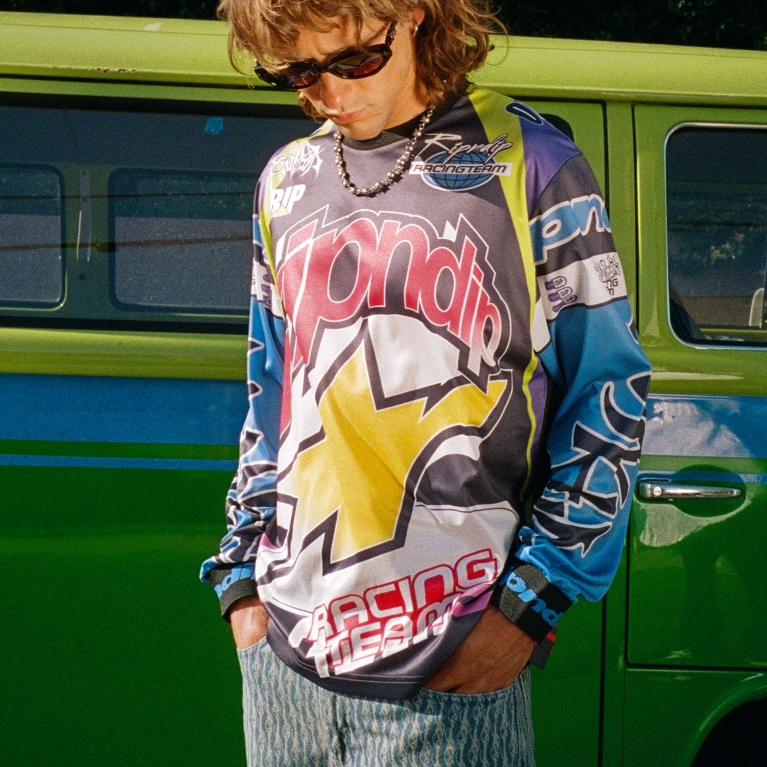 Wearing sunglasses and a Ripndip hoodie over the Allstar Racing Jersey - Multi-colour, a long-haired person in patterned shorts stands before a green van, looking down with hands in pockets. Gate21.ee
