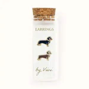 A clear glass phial with a cork stopper holds two dachshund-shaped earrings, one black and one brown, displayed upright. The phial is labelled "Tennis earrings by Vivi (Copy)" in gold lettering. Gate21.ee