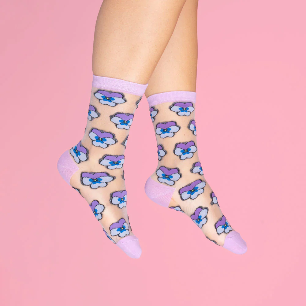A pair of feet wearing Coucou Suzette's Pansies - Transparent Socks, featuring sheer fabric with purple trim, heel, and toe, and decorated with a repeating blue and purple pansy pattern on a solid pink background. Gate21.ee