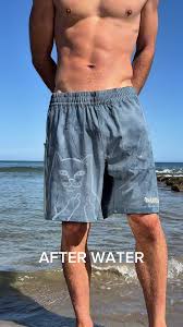 A person stands on a beach wearing Nermal Swim Shorts - Slate by Ripndip, featuring a faint cat design that appears when wet. The words AFTER WATER are written across the lower part of the image. Gate21.ee