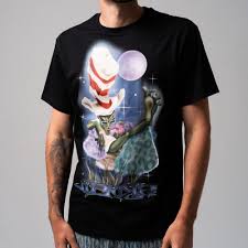 A person wears the black Here All Along Tee, which features an illustrated green cat in a red and white striped hat standing on a rock with a purple moon and starry background. The wearer’s face is not shown. Gate21.ee