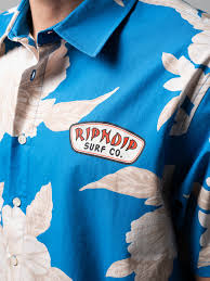A close-up of a person wearing a blue and white floral button-up shirt with a patch that reads RIPNDIP SURF CO. on the left chest. Gate21.ee
