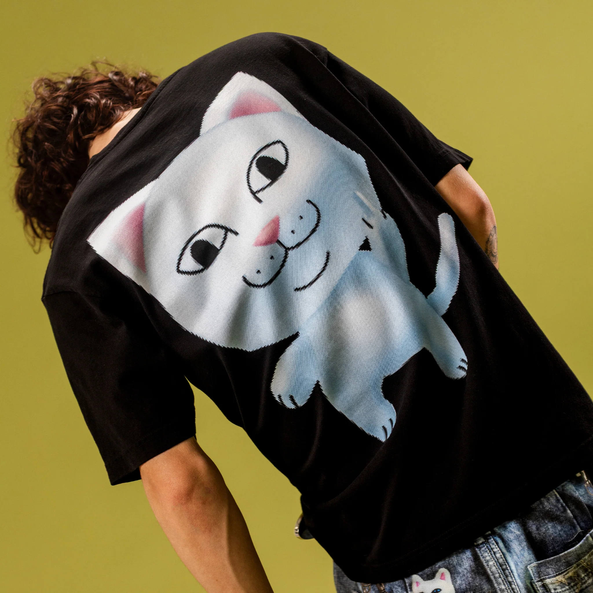A person with curly hair faces away, wearing the black Big Poe T-shirt featuring a large cartoon white cat with pink ears and a raised paw. The background is solid olive green. Gate21.ee