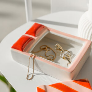 A ceramic jewellery box in the shape of a duck, inspired by the dreamy cat jewellery plate by Hello Ferretti, features red and pink accents and holds gold rings, a bracelet, and a necklace. The jewellery box sits on a white surface in bright natural light. Gate21.ee