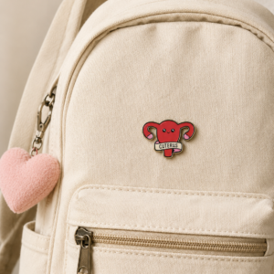 A beige rucksack with a pink heart keyring and a red “Cuterus - Pin” enamel badge shaped like a uterus attached to the front pocket. Next to it is a playful “This is fine” enamel badge, adding an extra touch of whimsy. The zip is closed, and the background is neutral. Gate21.ee