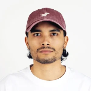A person with medium-length dark hair, light facial hair, and hoop earrings is seen wearing a green Moomin Cap featuring a white animal logo, paired with a plain white shirt as they look directly at the camera against a simple background. Adding a subtle touch of personality to their minimal look, a small Moomin keyring is attached to one of the hoop earrings. Gate21.ee