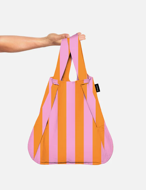 A hand holds the Stripes - Orange Notabag, a reusable shopping bag with vertical pink and orange stripes, against a plain white background. Gate21.ee