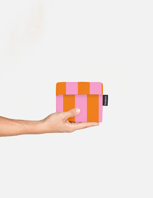 A left hand holds a small, square Notabag pouch from the Playground - Special Edition (Copy) collection, featuring vertical pink and orange stripes. The pouch has a black tag with white text on the right side, set against a plain white background. Gate21.ee