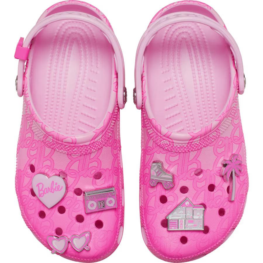 The Crocs Barbie® Classic Clog is a pair of pink Crocs featuring Barbie-themed decorations, such as heart, radio, car, flamingo, house, and heart-shaped sunglasses charms on the tops. The shoes are adorned with a repeating Barbie logo pattern. Gate21.ee