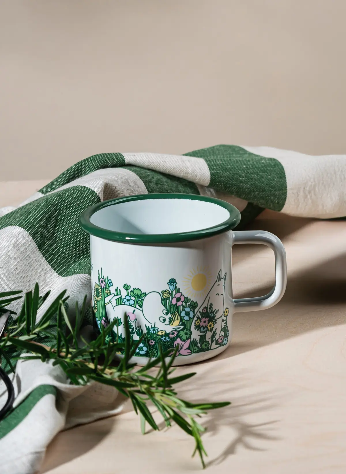 A pink enamel mug featuring Little My from Moomin, with a 3.7dl capacity, is placed on a light wood surface alongside a sprig of rosemary and a green-and-white tea towel in the background. Gate21.ee