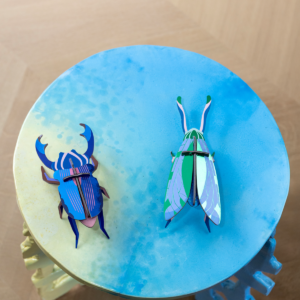 Two colourful, geometric Azure Stag Beetle paper models from StudioROOF are placed side by side on a round, blue and yellow gradient table with a pale wood floor in the background. Gate21.ee