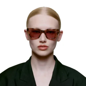 A person with fair skin and blond hair pulled back, wearing stylish KAWS - Black/Yellow Tortoise (Copy) sunglasses and a black blazer, faces the camera against a white background. Gate21.ee