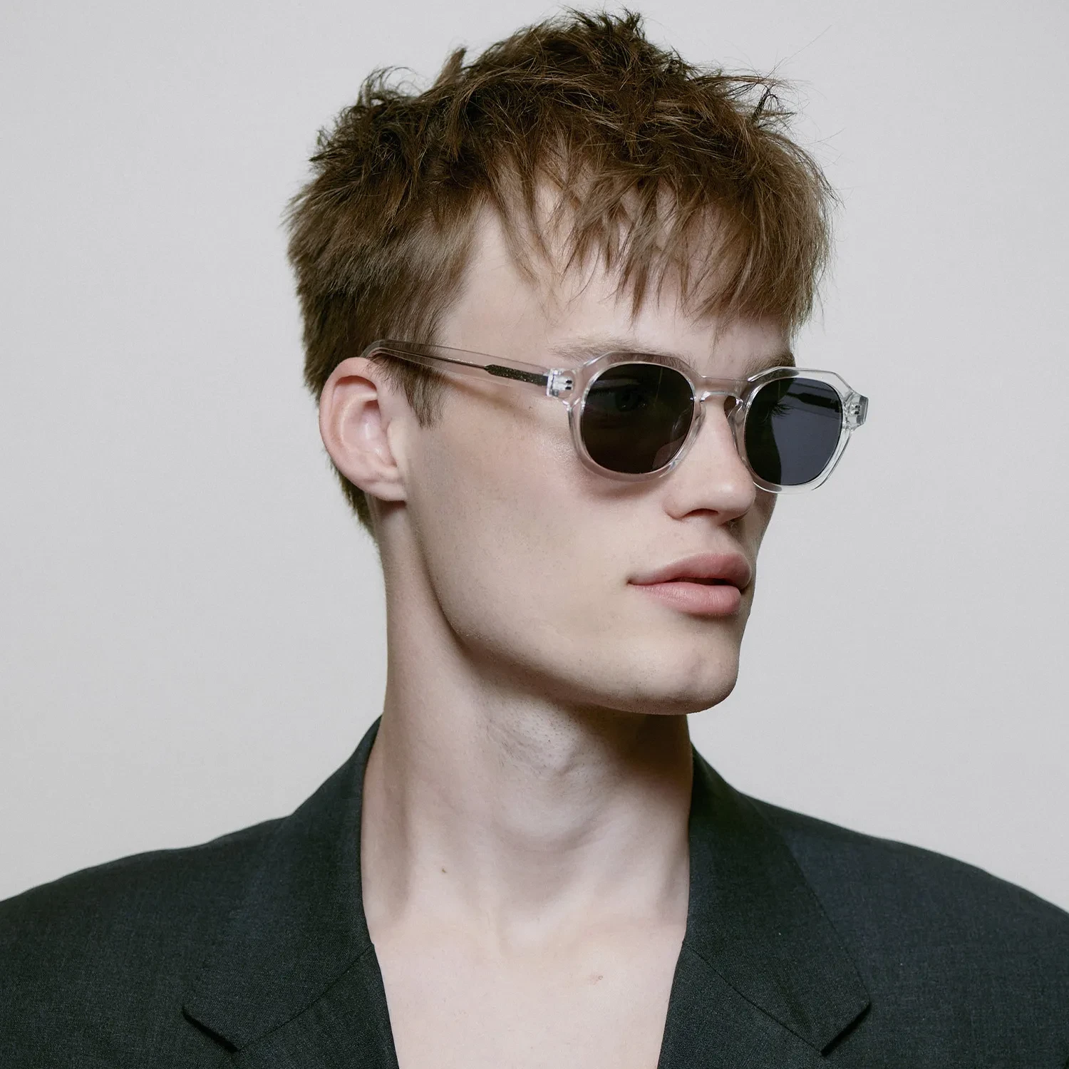 Young person with short, tousled brown hair wearing Zan - Crystal clear-frame sunglasses by A. Kjærbede and a dark blazer, facing slightly to the side against a plain light background. Gate21.ee