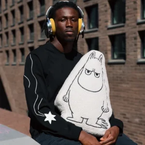 A person wearing yellow headphones and a black sweatshirt with a star on the sleeve sits outdoors, holding a Moomintroll Temper Cushion—a plush toy featuring the white, pointy-eared character—with a brick building in the background. Gate21.ee