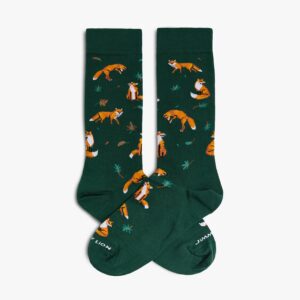 A pair of Dusk Fox - Green socks by Jimmy Lion, featuring a repeating pattern of orange foxes in various poses and scattered leaves on a dark green background. These crew-length socks are shown with the toes crossed over each other. Gate21.ee