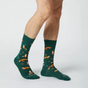 A person stands barefoot except for a pair of Dusk Fox - Green socks, featuring an orange fox pattern, against a white background. Only their lower legs and feet are visible. Gate21.ee