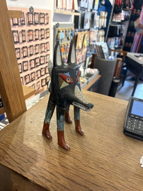 A painted wooden figurine, “Kauri hunt - suur,” with exaggerated wolf features and a sharp, legendary gaze stands on a wooden counter beside a payment terminal in a shop filled with diverse merchandise. Gate21.ee