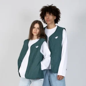 Two people pose together wearing matching Moomin Classics Vest Unisex - Green over white long-sleeved shirts and light blue jeans, evoking the playful charm of Moomin apparel. Set against a plain light grey background, each green quilted vest features a small white embroidered bird on the chest. Gate21.ee