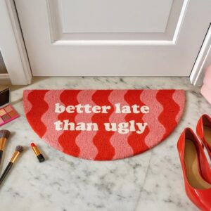 A semicircular “Better Late than Ugly” doormat, featuring red and pink wavy stripes with white lettering, is placed on a marble floor. Surrounding it are red high heels, make-up brushes, lipstick, a make-up palette, and a chic handheld fan—perfect for staying cool whilst you get ready. Gate21.ee
