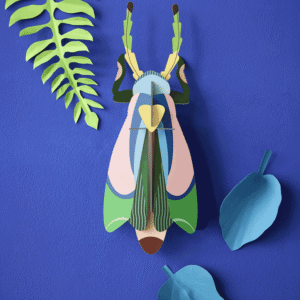 The Sakura Beetle, a vibrant paper sculpture by StudioROOF, is showcased on a blue background and surrounded by three green and blue paper leaves. Gate21.ee