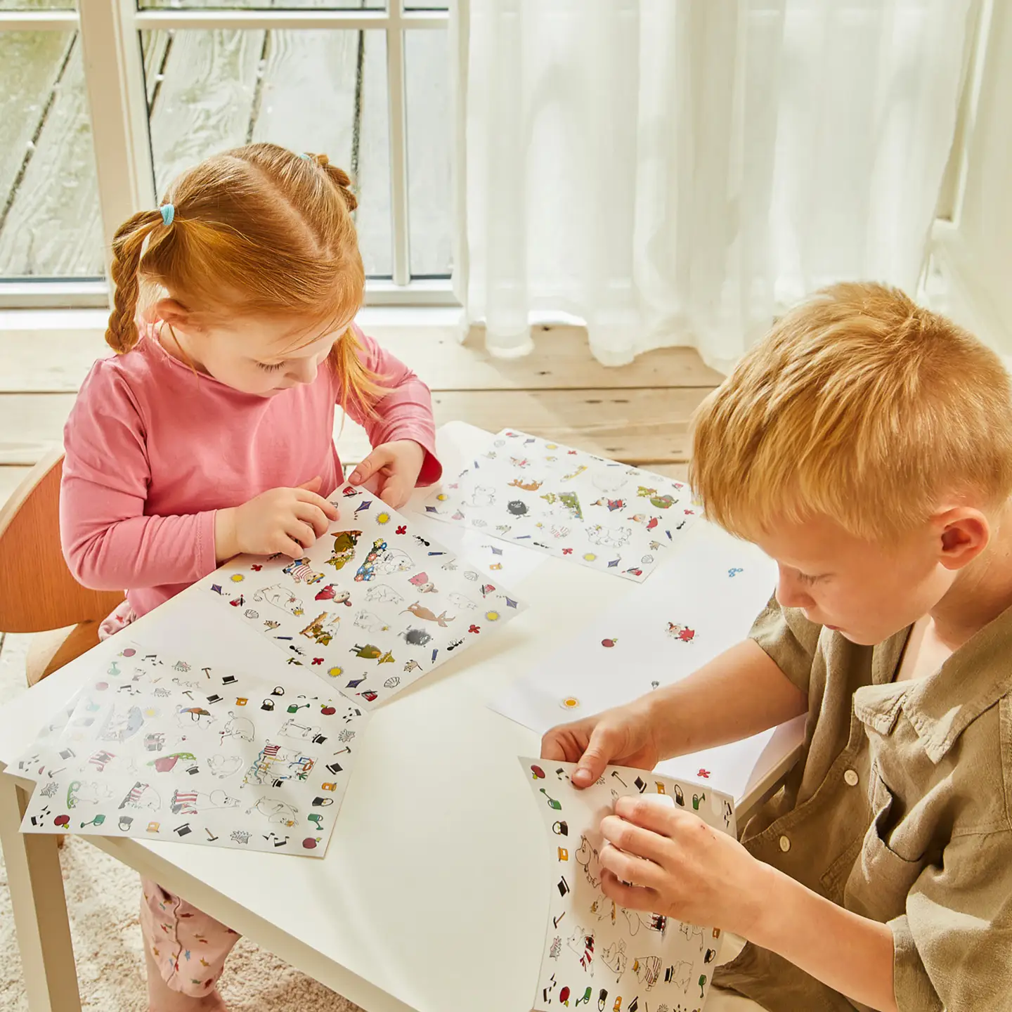 A young girl and boy sit at a white table by a window, each holding sheets of Moomin - Stickers - Moominvalley. They focus on peeling and placing the colourful stickers, with sunlight streaming through sheer curtains in the background. Gate21.ee