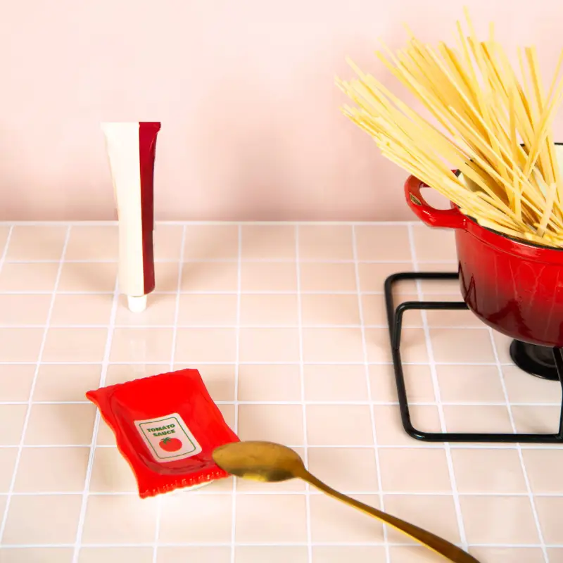A red “Ketchup - Plate” pot holding uncooked spaghetti sits on a stand, evoking the artistry of L’expressionist gifts. Nearby, a gold spoon, a small red packet labelled tomato, and a capped tube are neatly arranged on a tiled counter against a pale pink wall in the background. Gate21.ee
