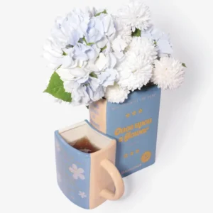 A light blue book-shaped vase with white and light blue flowers stands next to the Morning Story - Mug, which is filled with tea. The vase reads “Once upon a flower,” and both items, perfect as L’expressionist gifts, feature delicate white floral designs. Gate21.ee
