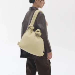 A person with dark hair in a low bun wears a brown jacket and trousers, carrying the large, cream-coloured ÖLEND Ona Soft Bag - Chocolate with knot details over their shoulder. The background is plain and white. Gate21.ee