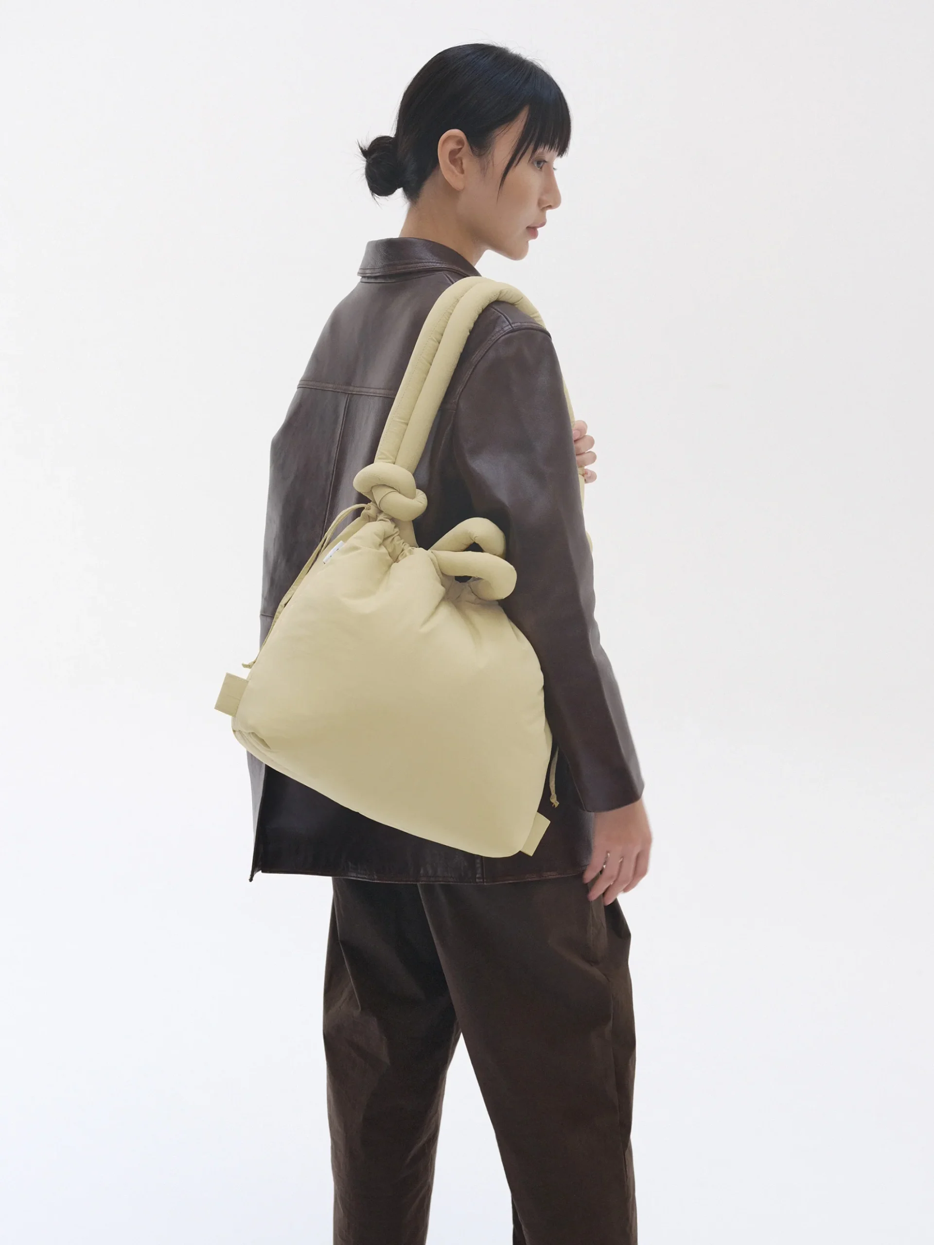 A person with dark hair in a low bun wears a brown jacket and trousers, carrying the large, cream-coloured ÖLEND Ona Soft Bag - Chocolate with knot details over their shoulder. The background is plain and white. Gate21.ee