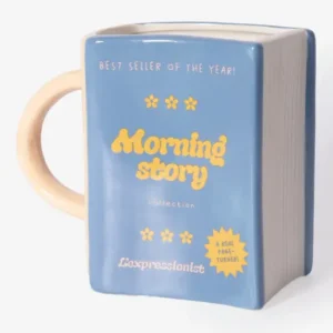 Introducing the Morning Story - Mug: a blue ceramic mug designed in the shape of a closed book, featuring a cream-coloured handle and edges. The front displays “Morning Story” in yellow text, accented with decorative flowers and additional white and yellow lettering. This unique piece is an ideal addition to any L’expressionist gift collection—perfect for book lovers who appreciate artistic flair. Gate21.ee