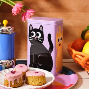 A Kitchen Storage / Coffee Tin - Cats with a purple wrap featuring a cartoon black cat sits next to a plate of small cakes with pink icing, a blue cafetière, a vase with pink flowers, and a bowl of fruit, all arranged on a colourful surface. Gate21.ee