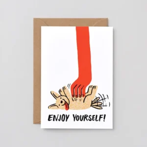 Introducing the “Enjoy! - Greeting Card”: a greetings card adorned with an illustration of a joyful dog lying on its back, delighting in scratches from a large red hand. The message below reads ENJOY YOURSELF! The “Enjoy! - Greeting Card” is displayed atop a brown envelope. Gate21.ee