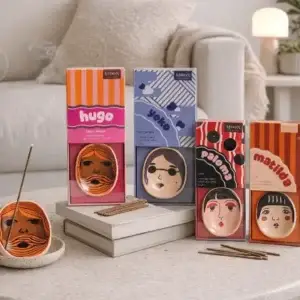 Five boxes of Yoko Faces - Incense Burner products from Wick & Vessel home fragrance, each featuring illustrated faces and names on the packaging, are arranged on a table. One incense coil is burning in a holder, with several sticks placed nearby. A sofa and decor can be seen in the background. Gate21.ee