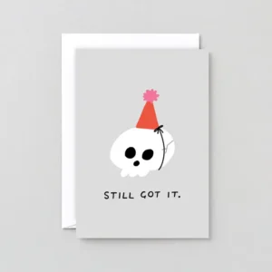 The Still got It! - Greeting Card features a cartoon skull in a red party hat with a pink pom-pom and bold “STILL GOT IT” text, set against a plain light grey background. Gate21.ee