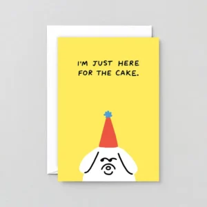 A yellow greeting card titled “Just here for Cake!” features a simple illustration of a dog in a red party hat with a blue pom-pom and the phrase “I’M JUST HERE FOR THE CAKE.” A white envelope is partially visible behind the card. Gate21.ee