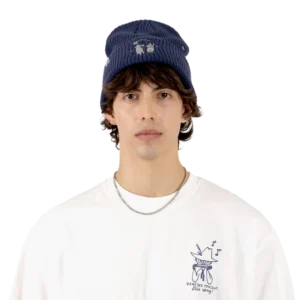 A young person with medium-length brown hair wears the Moomin Beanie in navy, a white graphic t-shirt with a drawing and text, and a silver chain necklace adorned with Moomin keyrings. The background is plain white. Gate21.ee